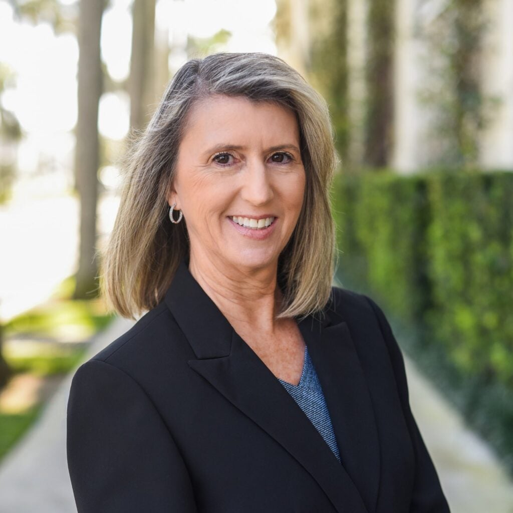 Smiling woman in blazer with blurred greenery background.