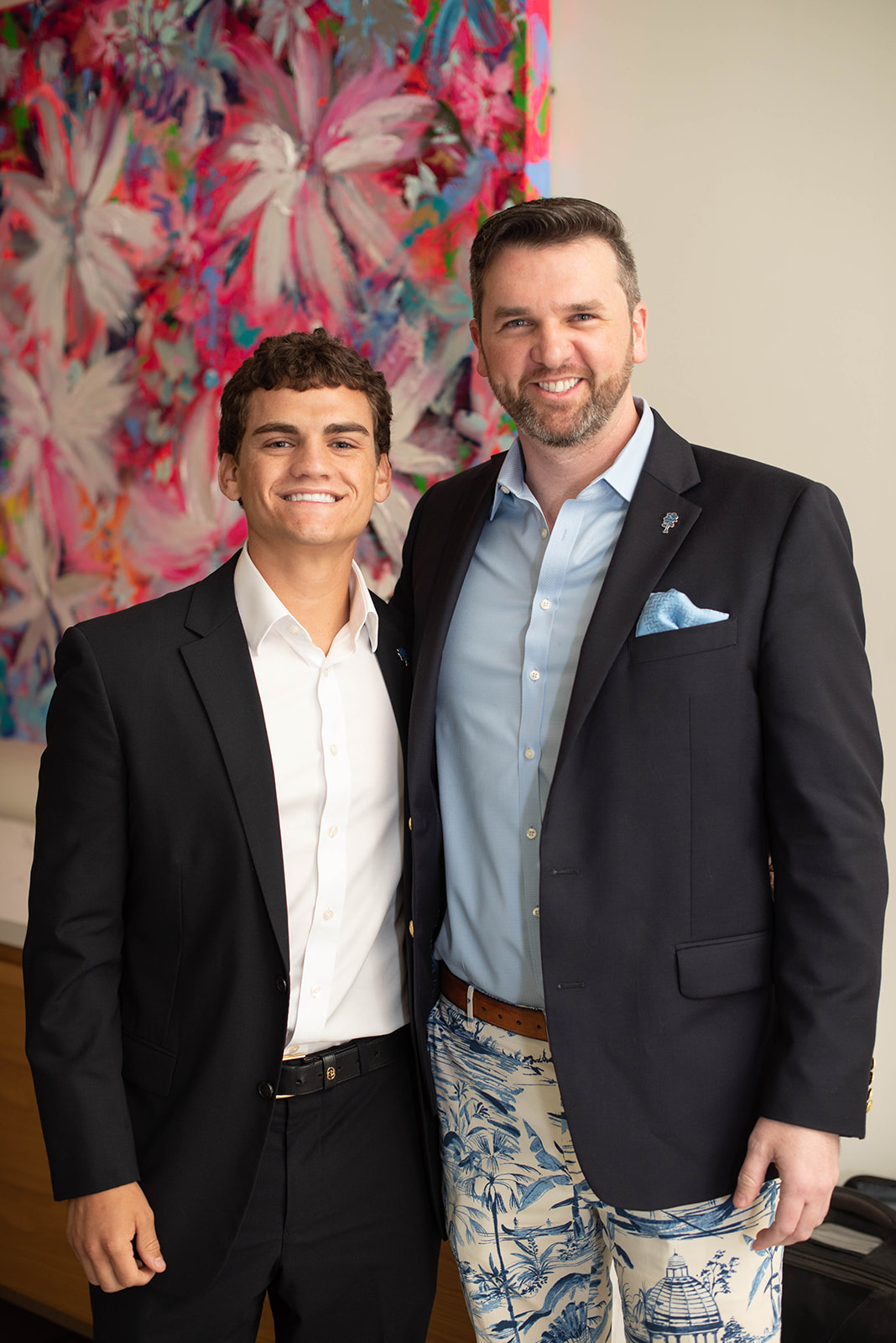 Dos hombres sonriendo con blazers posan frente a una obra de arte colorida.