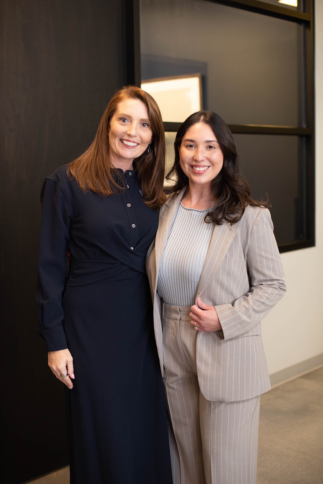 Dos mujeres sonriendo posan en un pasillo de oficina moderno.